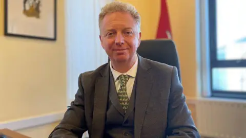 BBC Alex Allinson sitting at his office desk with a standing Isle of Man flag in the background. He has short, fair, wavy hair and is wearing a grey suit and waistcoat with a green patterned tie and white shirt.