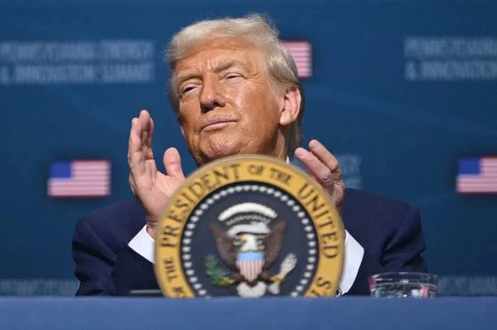 A man in a suit claps his hands at a podium with the U.S. presidential seal, against a backdrop with U.S. flags and text
