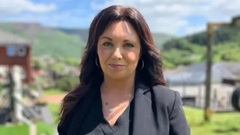 Leanne Gough stands outside with homes and hills behind her.