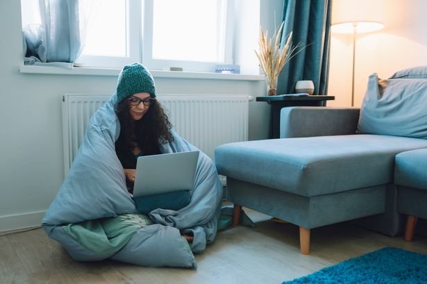 Middle age business woman work at home sitting on a floor next to heater wrapped in blanket and wearing woollen hat