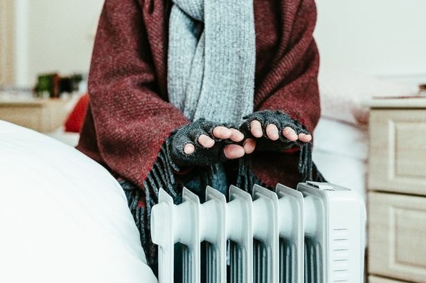 A retired senior man in his 70s sits at home inside his cold house in winter. It is so cold that he is wrapped up in warm winter clothing, including gloves and blanket, and is holding his hands over an electric heater for some extra warmth and comfort.