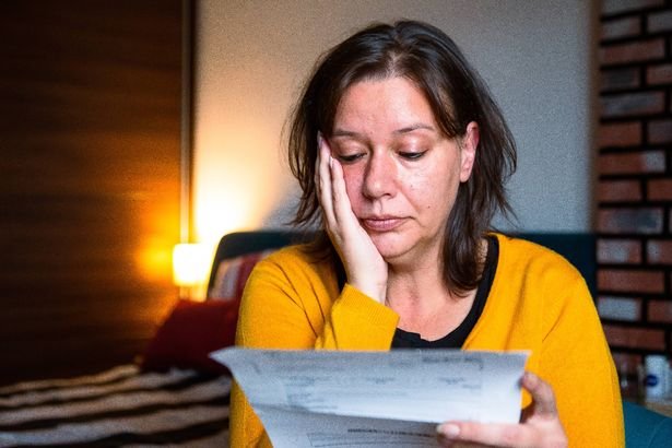Portrait of a mid adult woman checking her energy bills at home, sitting in her bedroom. She has a worried expression and touches her face with her hand while looking at the bills.
