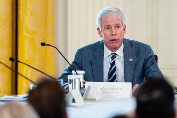 A man in a suit speaks at a conference, seated at a panel table with a name card