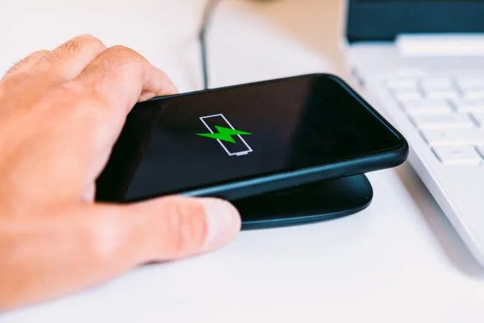 A hand places a phone on a wireless charger next to a laptop, with a battery icon showing charging progress