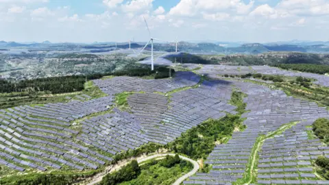 Getty Images An aerial view of a photovoltaic solar farm sprawling across undulating green hills in Shandong Province, China. The are also several white wind turbines rising in the background. A winding road curves through the lower foreground.