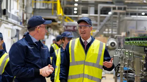 PA Media Peter Kyle being led around a glass factor in Cheshire - he is gesturing with his left hand, while standing among the machinery - he is wearing a baseball cap and a high visibility jacket 
