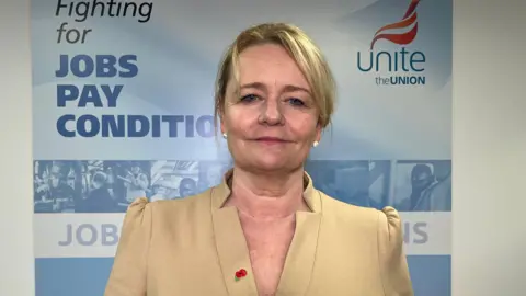 Sharon Graham standing in front of a banner with Unite's branding on it - she is wearing a cream / gold structured top,  has blond hair, is wearing pearl-like earrings, has a poppy on her lapel, and has a neutral expression 