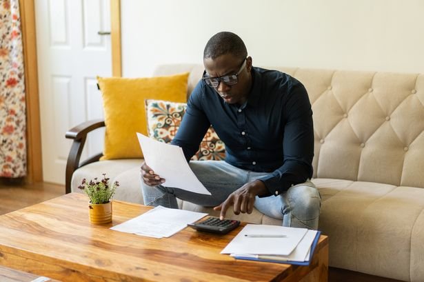 One man sitting on a sofa is holding a document while using a calculator in his living room