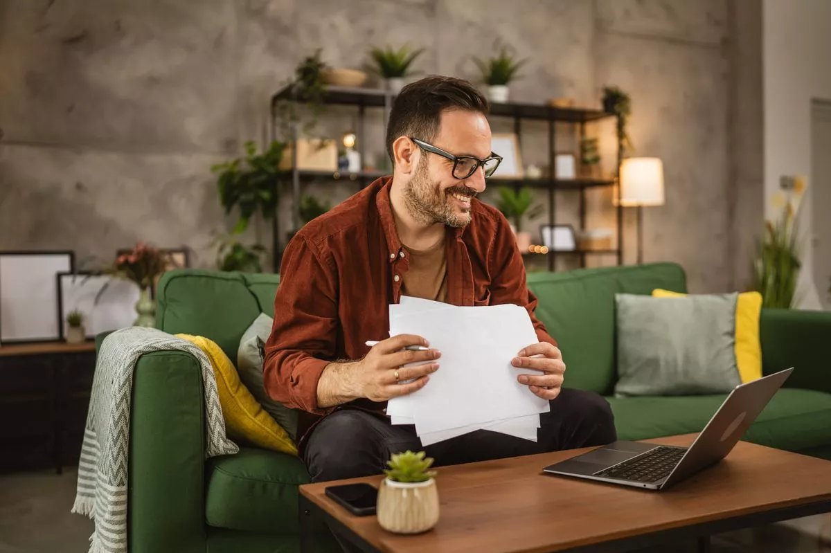 Adult caucasian man with eyeglasses work from home on laptop and hold documents