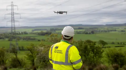 Scottish Power Utility worker in high-visibility clothing operating a drone near overhead power lines in a rural landscape, with pylons and green fields in the background