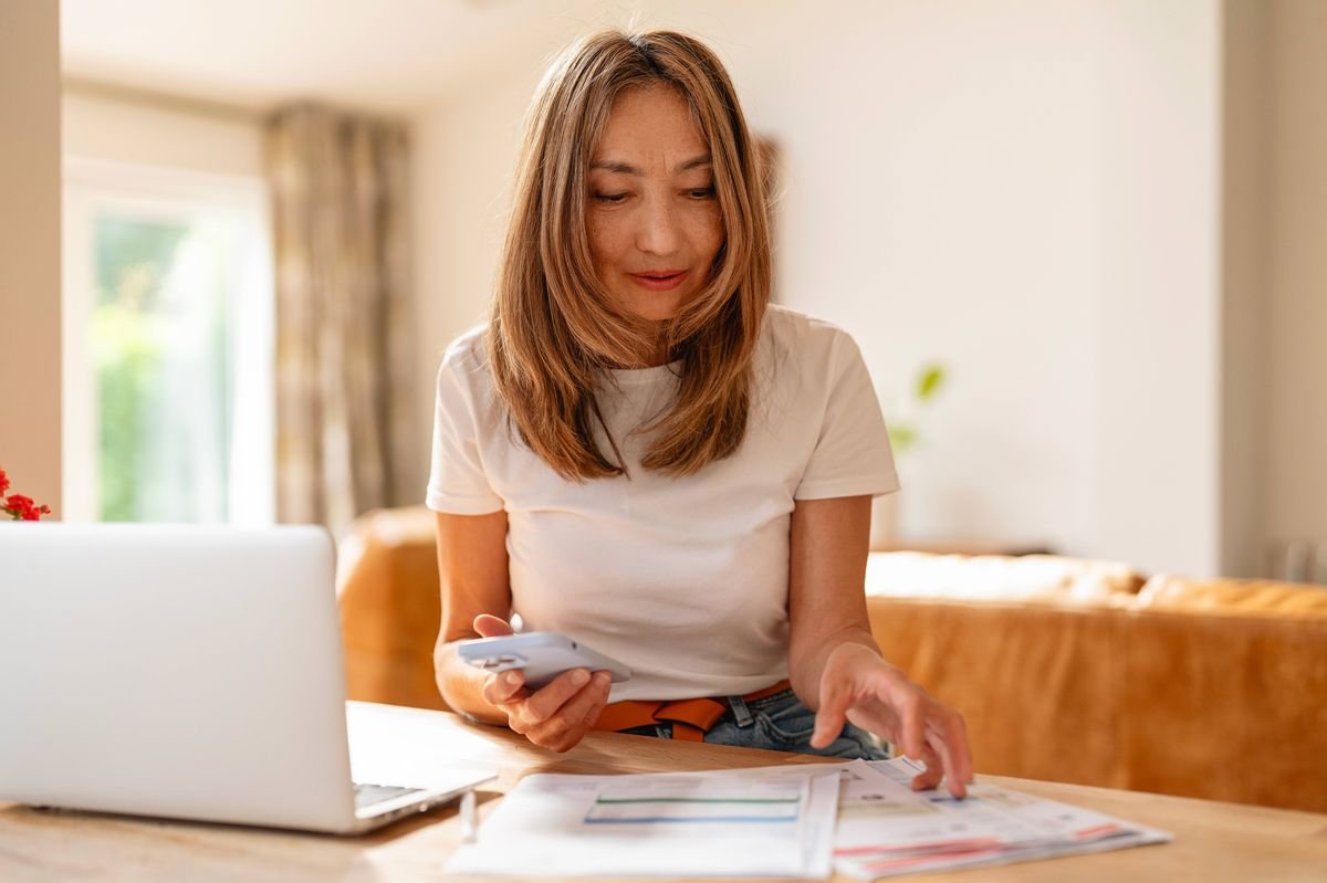 A woman is sitting at a wooden table, holding a smartphone and sorting through paper bills. 