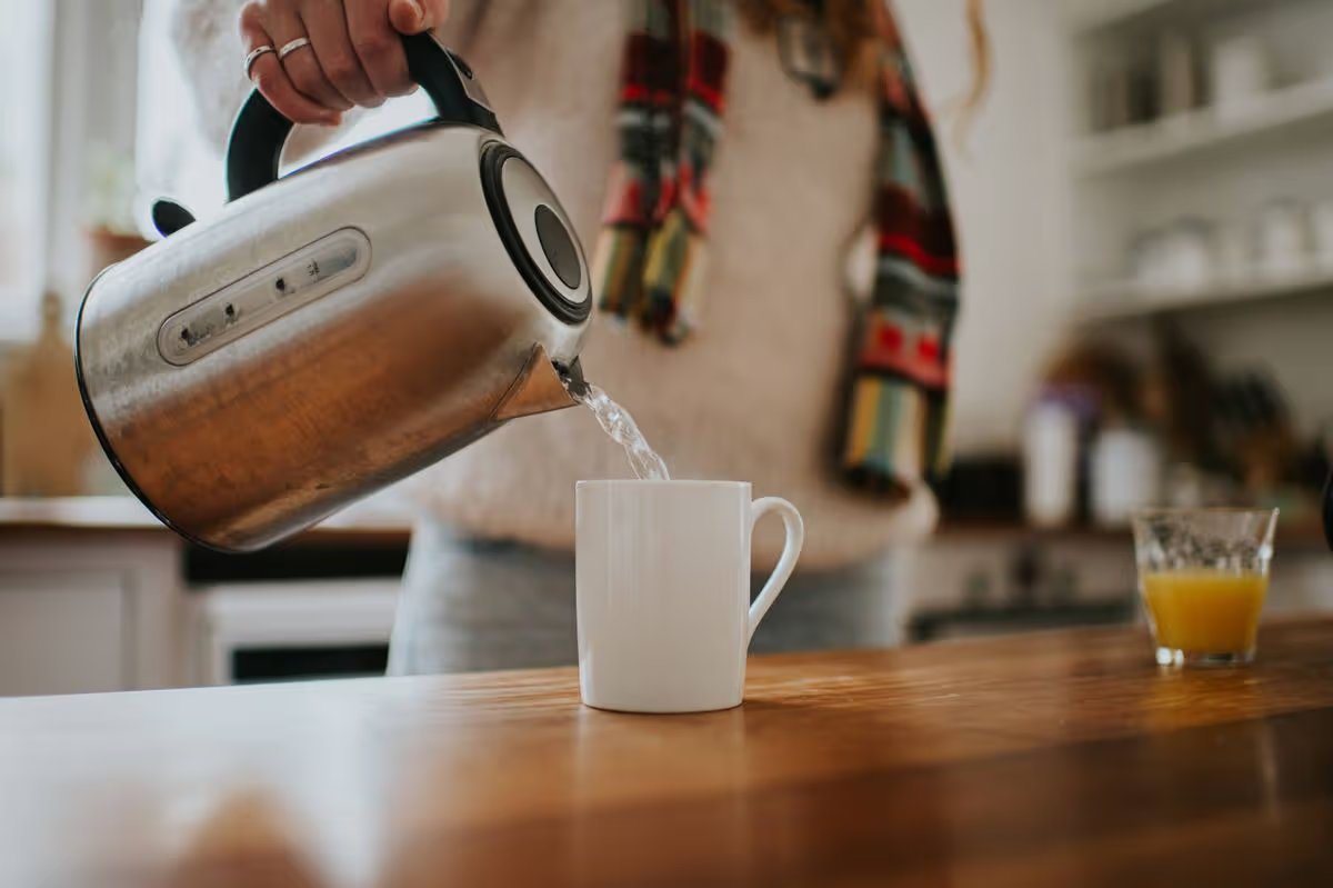 A woman pours boiling water into a white mug from a silver kettle