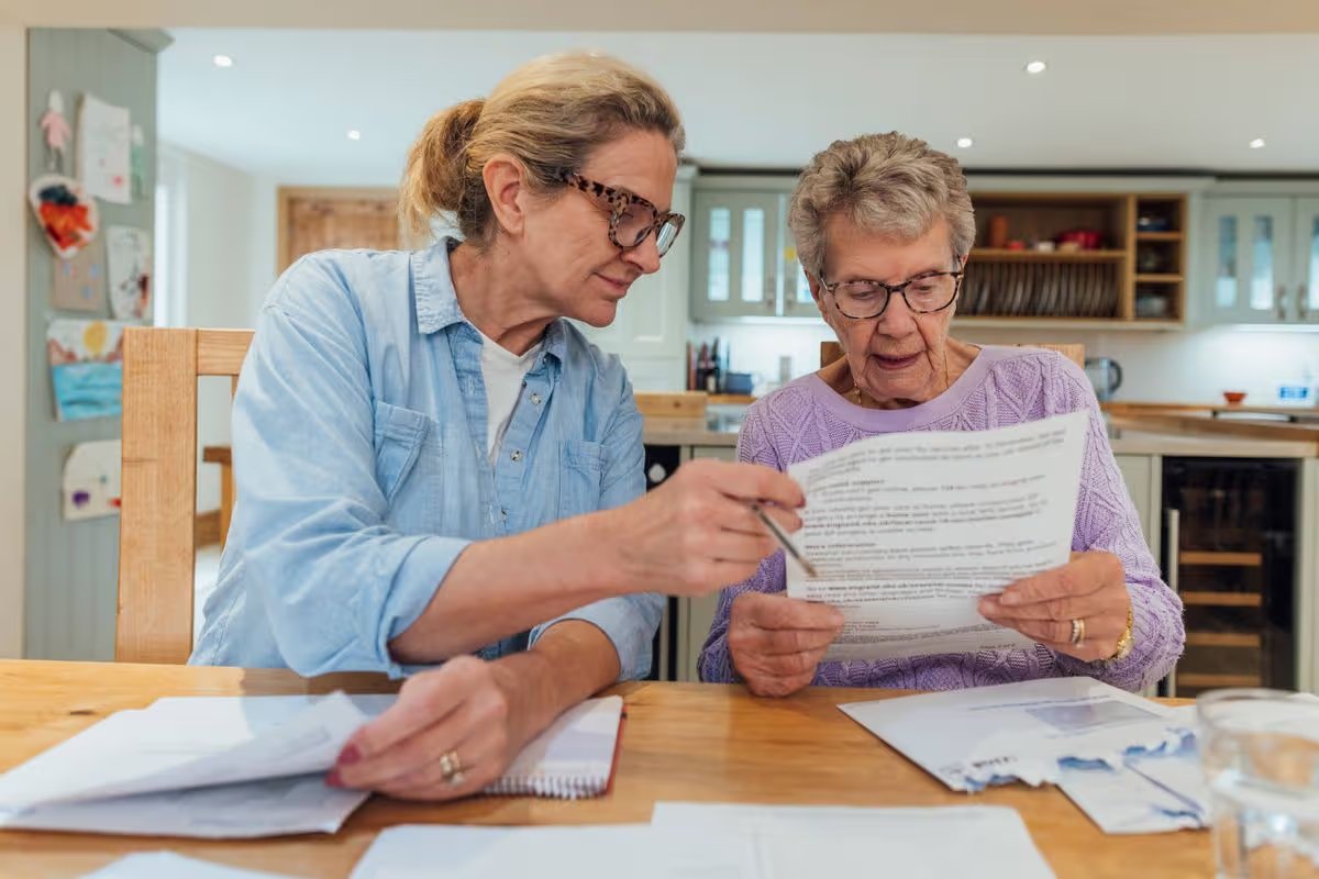 A senior woman and her mature daughter at home in Seghill, Northumberland. The senior woman is staying with her daughter for support and care as she has dementia. They are sat at the dining room table where they're looking through financial bills, looking concerned.