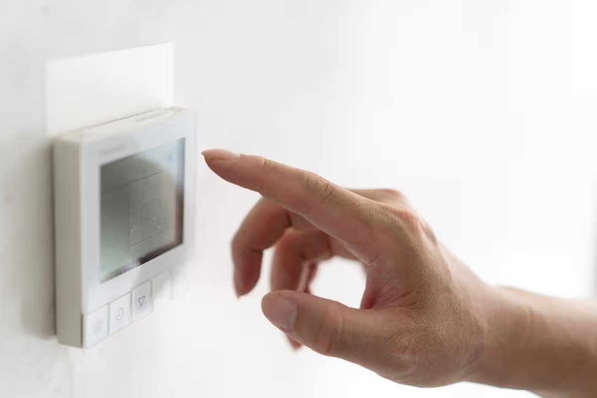 Hand adjusting digital thermostat on white wall, close-up.