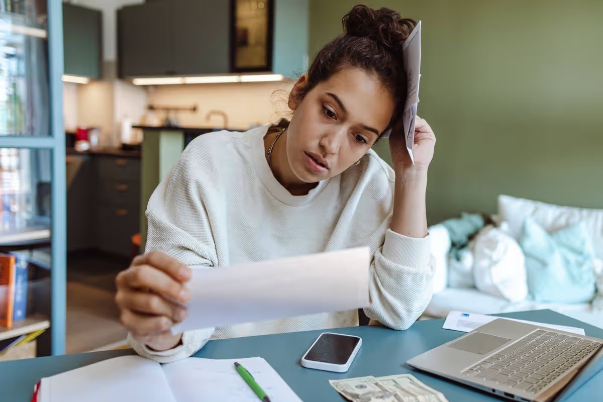 Young woman in a beige sweater sitting at the table and going through paperwork.  She is holding unpaid bills