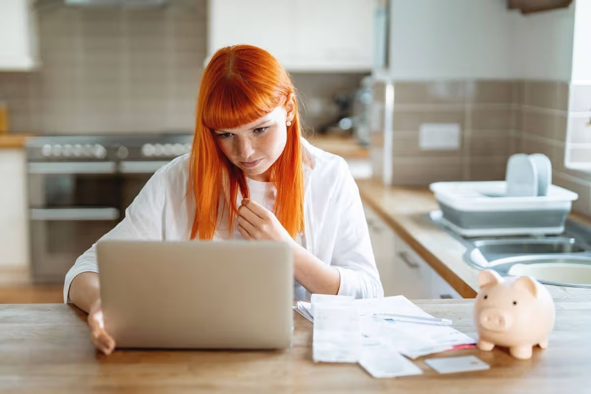 Young woman working on laptop while reviewing bills in home kitchen during daytime