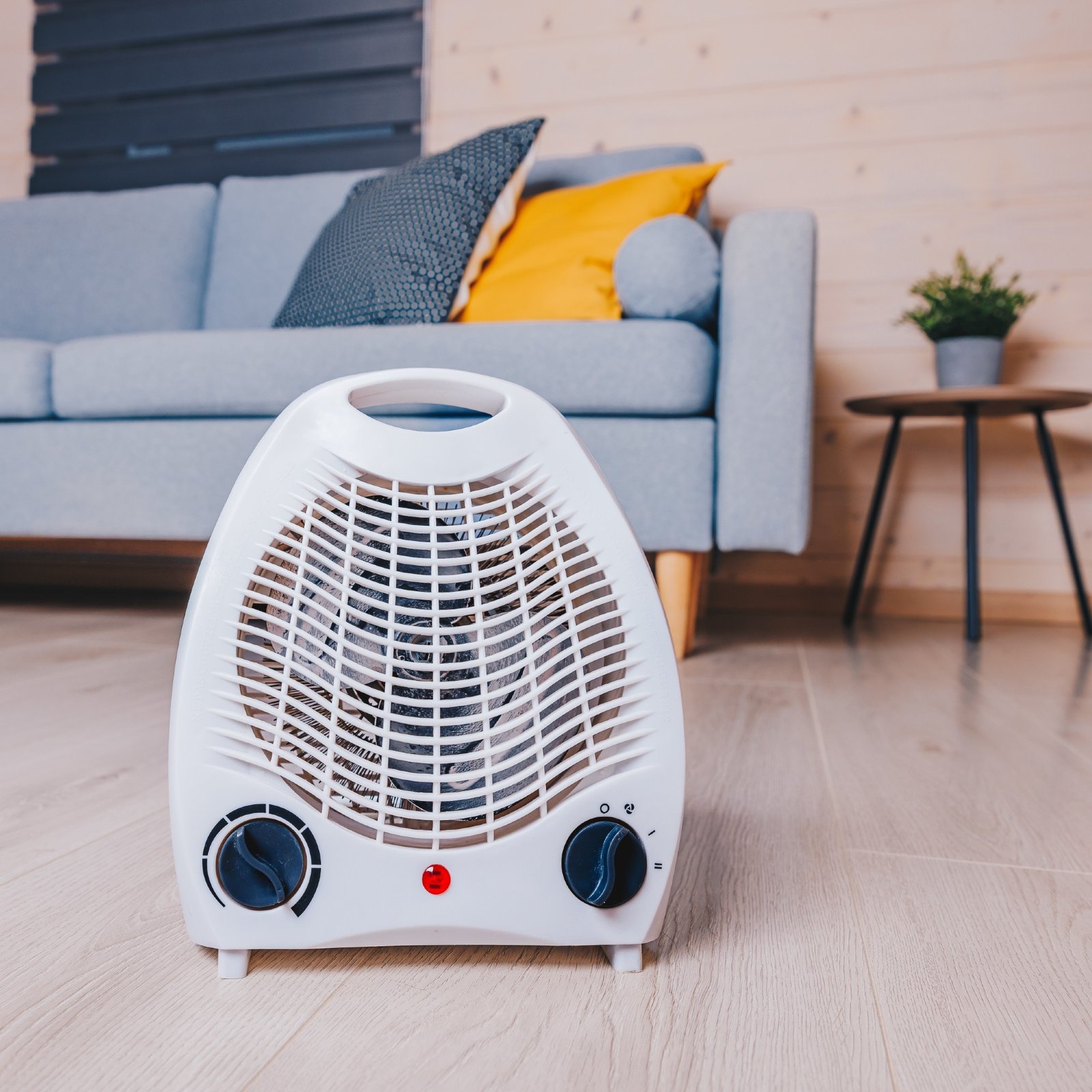 A small electric fan heater on the floor in a living room in front of a light blue sofa with dark blue and yellow cushions