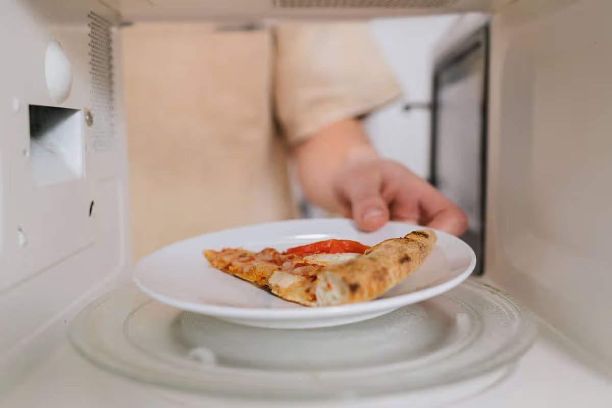 A person places a plate with a slice of pizza into a microwave oven for heating.
