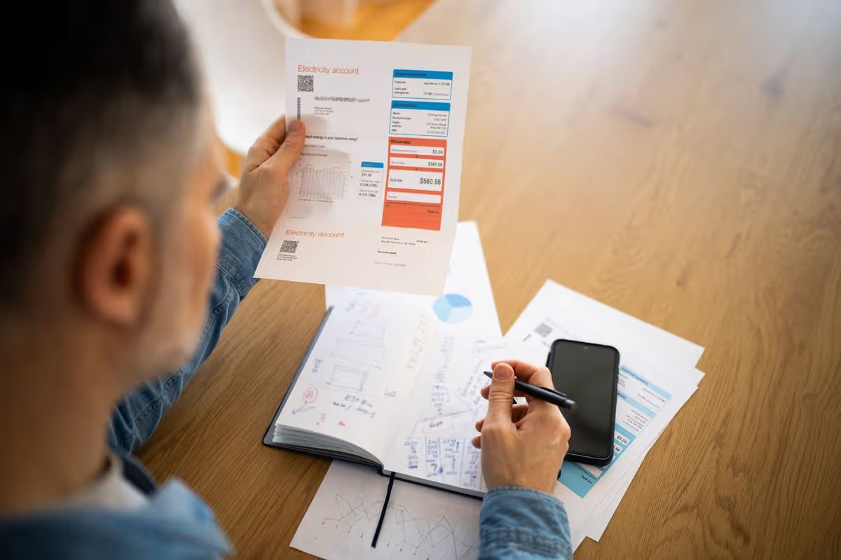 A man is sitting at a desk, holding an electricity bill in one hand and a smartphone in the other. Various financial documents and a notebook with sketches are spread out on the table, indicating a detailed review of expenses. The setting is bright and organized, reflecting a focused atmosphere.