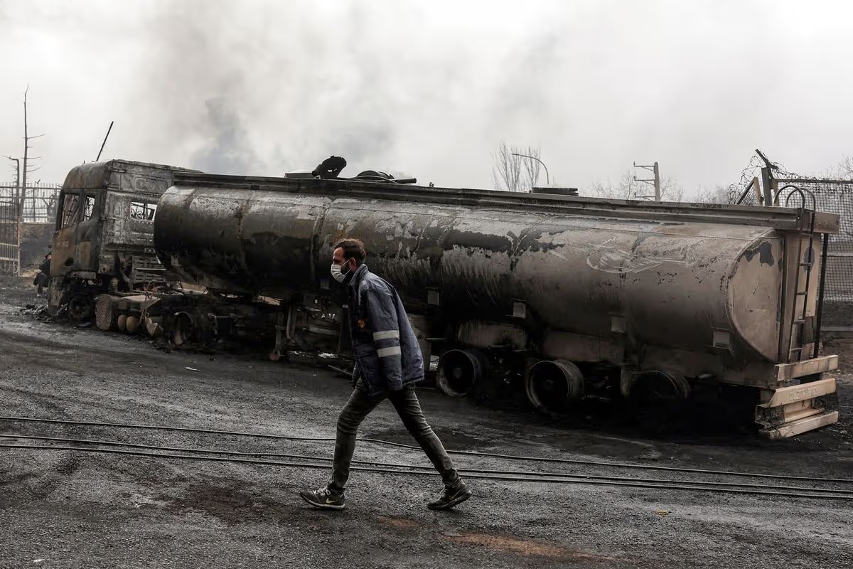 An Iranian civil defence member walks next to a destroyed fuel tanker vehicle near an ongoing fire following an overnight airstrike on the Shahran oil refinery