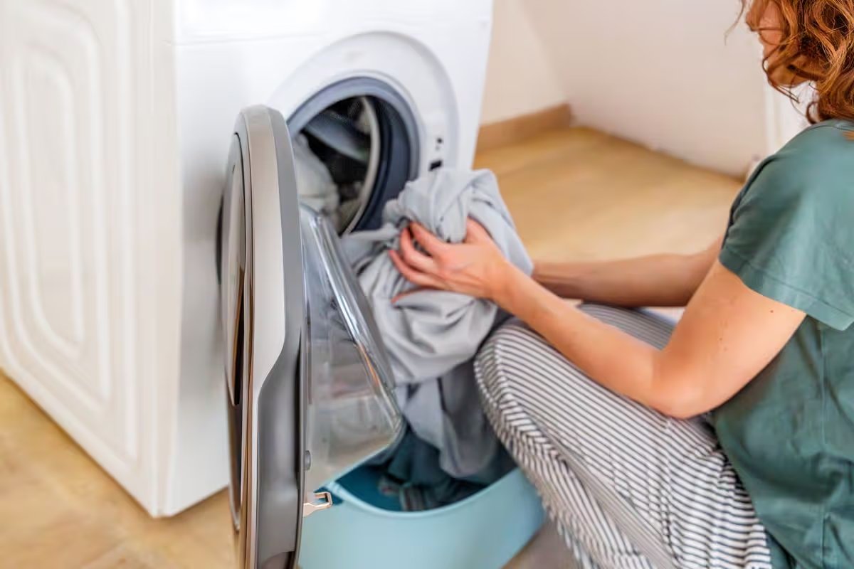 A cropped shot of a woman loading clothes into the drum of a white front-loading washing machine from the laundry basket