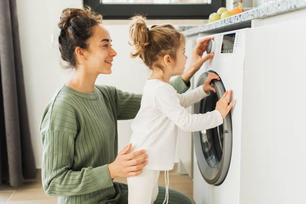 Mother a housewife with a baby engaged in laundry fold clothes into the washing machine. Beautiful mother and her daughter enjoying and laughing doing the laundry in the kitchen of modern apartment where a washing machine can be seen on.