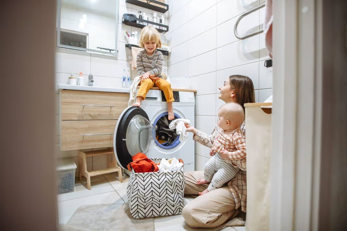 Mom multitasking laundry with two young children in cramped home bathroom