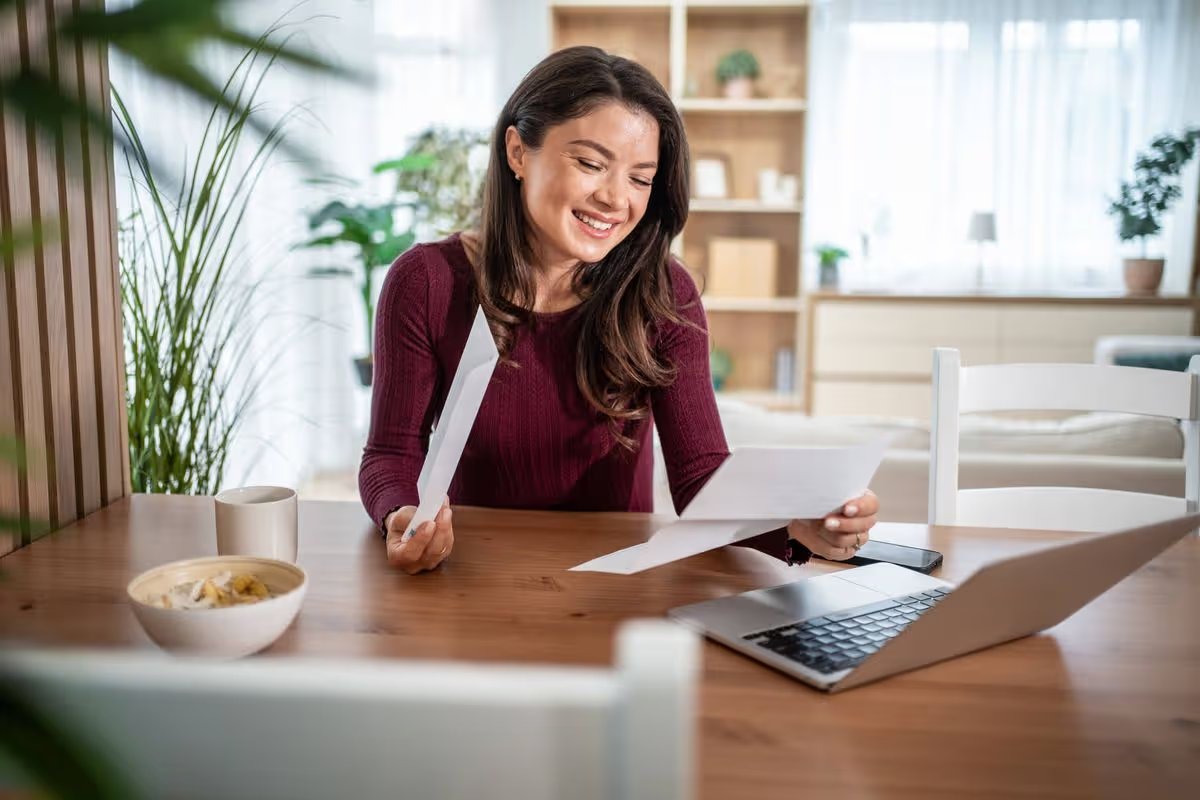 Young woman smiling as she reads an official letter at her kitchen table with laptop, celebrating good news about finances, application approval or business success at home
