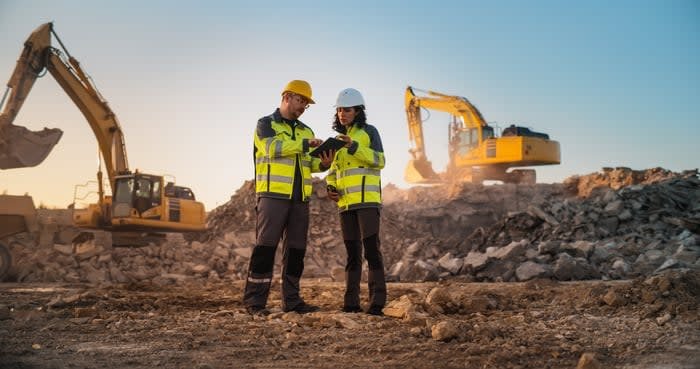 Two engineers in hardhats and yellow vests review a tablet while standing at a construction site in front of two excavators.