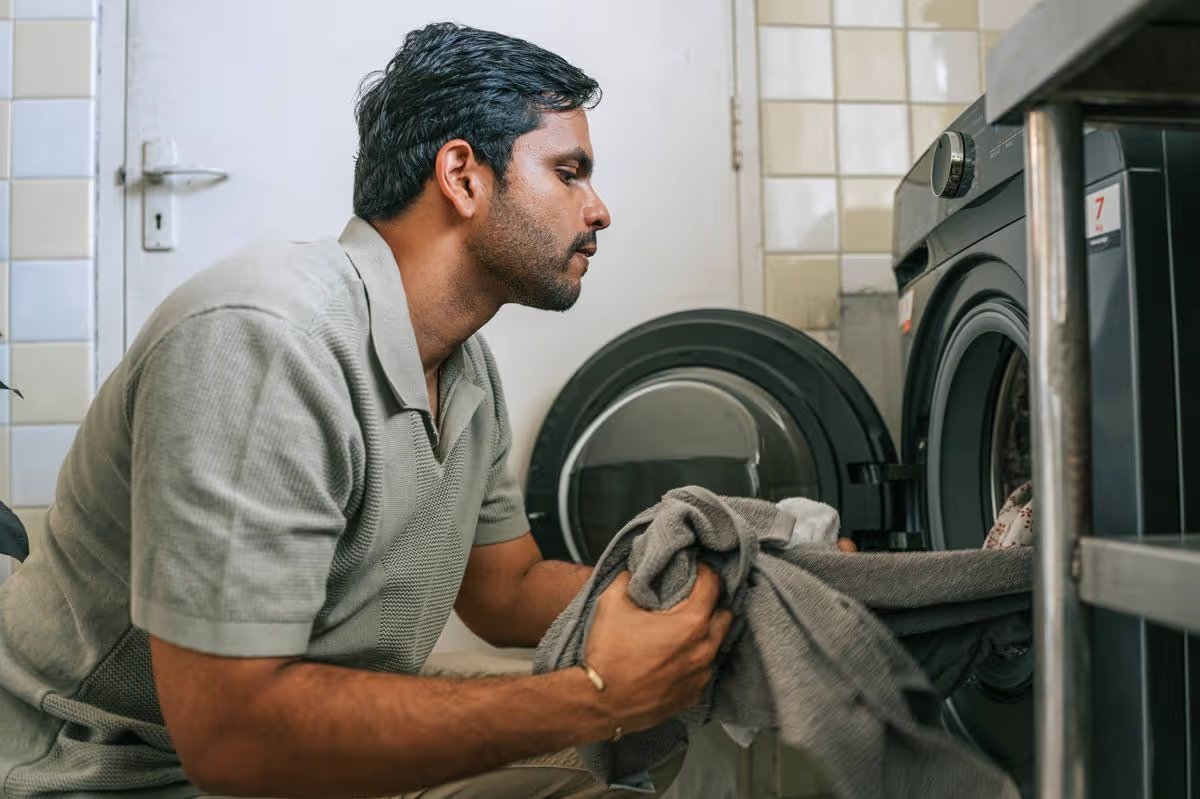 Asian man loading laundry into a front-loading washing machine in a home laundry room