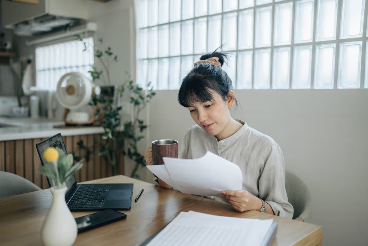 Young focused woman in spectacles calculating budget at home, sitting at table with laptop, holding paper taxes bills and reading financial information.