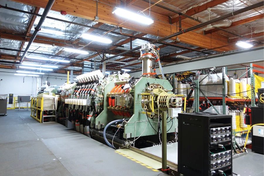 A long, pale green fusion prototype stretches across a bright industrial facility, with metal chambers, cables and control equipment along its length.