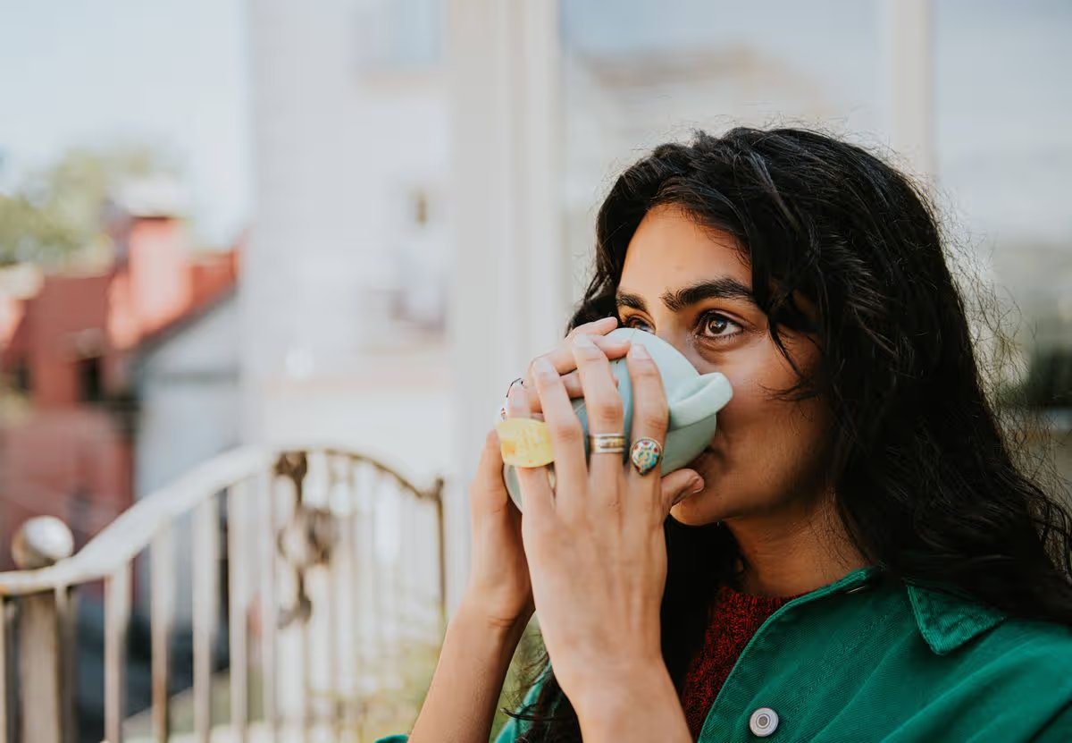 A woman cups her hands around a mug of herbal tea and drinks the brew.