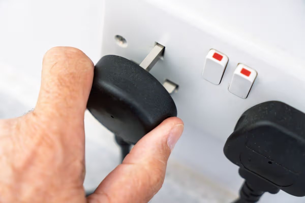 A close-up view shows a human hand gripping and inserting a black three-pin electrical plug attached to a power cord into a white wall-mounted electrical socket. 