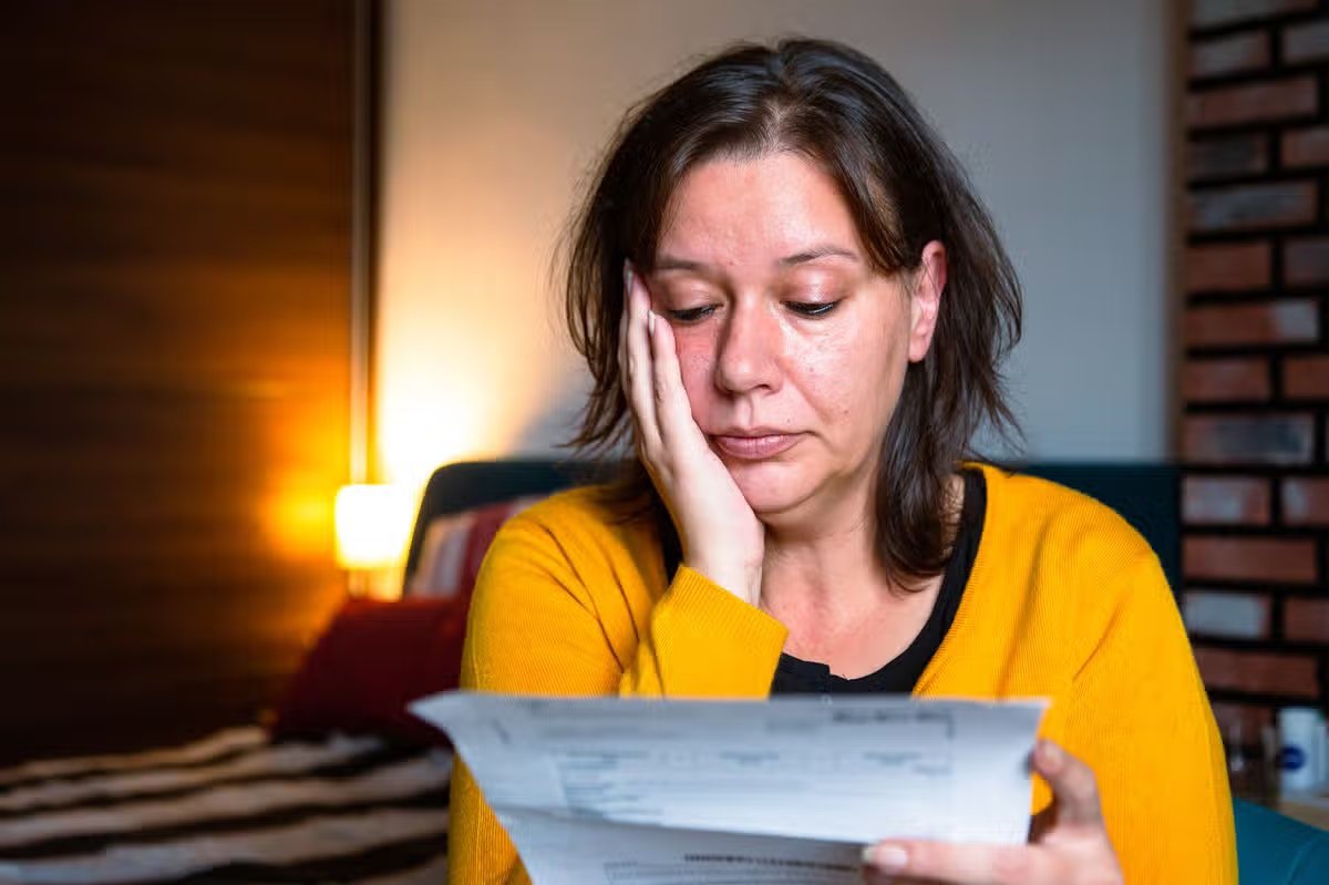 Portrait of a mid adult woman checking her energy bills at home, sitting in her bedroom. She has a worried expression and touches her face with her hand while looking at the bills. Focus on the woman while the interior architecture of the bedroom is defocused.