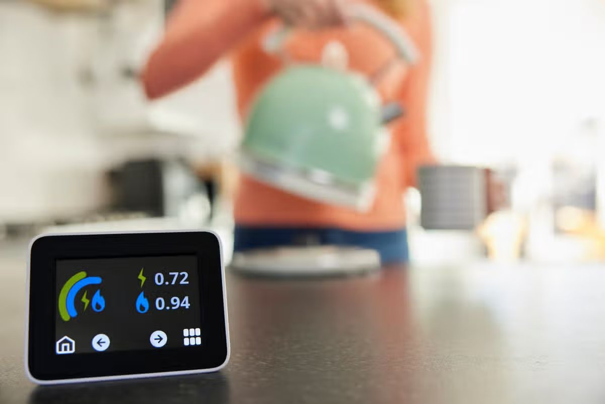 Close Up Of Smart Energy Meter In Kitchen Measuring Electricity And Gas Use With Woman Boiling Kettle.