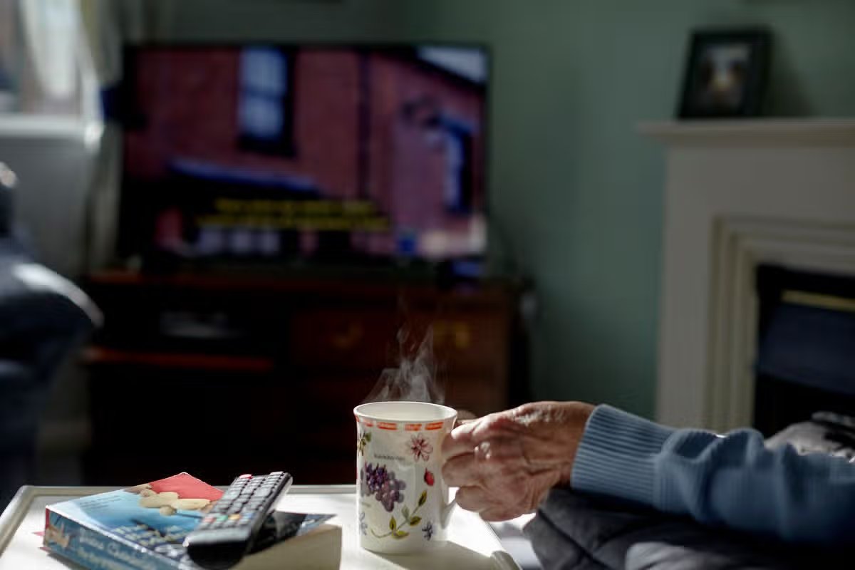  Picture shows an elderly hand holding a hot cup of tea with steam coming from it, TV on out of focus in the background, book and TV remote by her on the side table