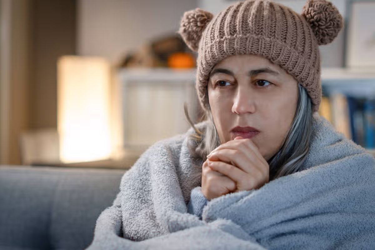 Woman struggling with cold temperatures at home, wrapped in a blanket and wearing a beanie